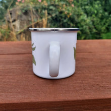 Load image into Gallery viewer, View of the handle, showing how the design wraps around the mug. Under the cup is a brown picnic bench, behind which is an overgrown wall.
