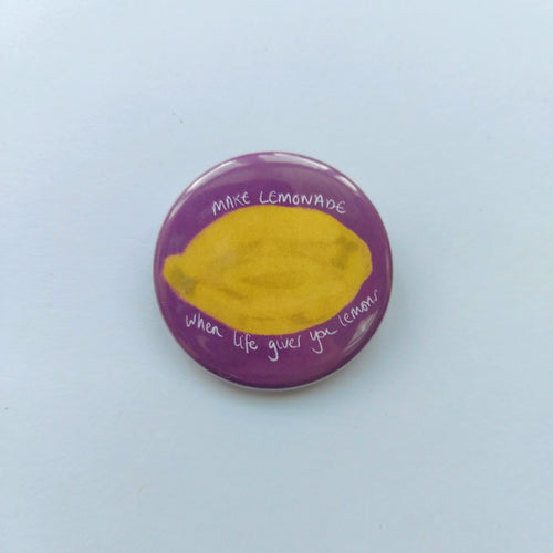 A pink badge sits on a white background. The badge features a yellow textured lemon with the words 'when life gives you lemons MAKE LEMONADE' in white.