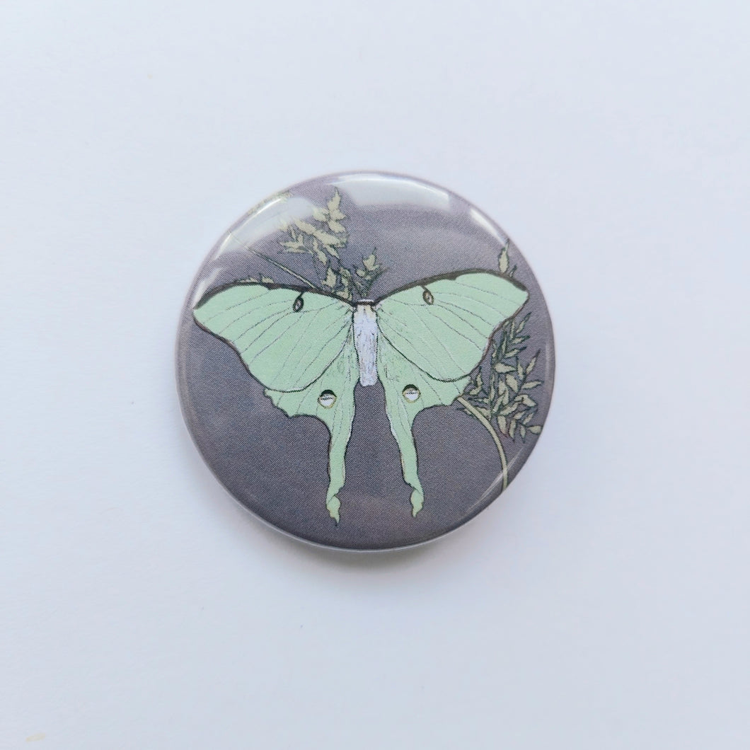 A lilac grey circle badge can be seen in the middle of the photo in front of a white background. The badge features a pastel green luna moth in the centre with a golden cow parsley behind it.
