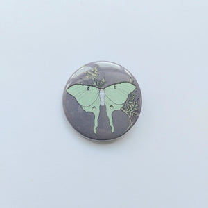 A lilac grey circle badge can be seen in the middle of the photo in front of a white background. The badge features a pastel green luna moth in the centre with a golden cow parsley behind it.