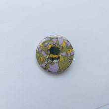 Load image into Gallery viewer, A lilac circle badge in front of a white background. The badge features a fluffy golden yellow, black and grey bee in front of yellow, white and green honeysuckle
