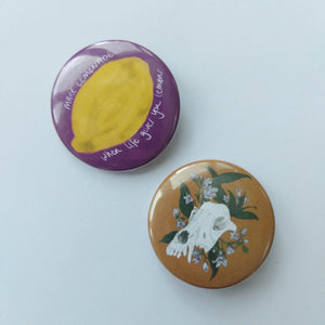 Two circle badges sit on a white background. The one on the top left is a pink badge featuring a yellow lemon with the words 'MAKE LEMONADE when life gives you lemons' written in white handwriting around it. Below and to the right of it is a mustard brown badge featuring a white dog skull with pale purple dogbane growing up around it. 