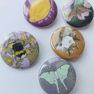 Five badges are visible across a white background, from L to R - a lilac badge with a fluffy bumblebee and honeysuckle across it, a pink badge with a yellow lemon and white writing. A lilac grey badge with a pastel green luna moth and golden cow parsley, a mustard brown badge with a dog skull and dogbane on it and a dark blue badge featuring an elephant hawk-moth in front of some pink evening primrose.