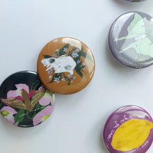 Load image into Gallery viewer, Four badges are visible scattered across the image from L to R top to bottom you can see a dark blue badge with a elephant hawk moth and pink evening primrose, a mustard brown badge featuring a dog skull and purple dogbane, a lilac grey badge with a pastel green luna moth and golden cow parsley and a pink badge with a yellow lemon and white writing on it.

