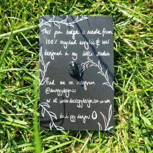 The back of the black backing card featuring information about the pin badges in white handwriting, as well as the outline of leafy vines. Behind the backing card is a green grassy background. 