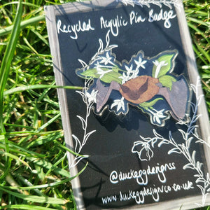 A close up of the pin badge featuring a brown and grey pipistrelle badge with night flowering jasmine bat. Behind the pin badge is a black backing card featuring a white design, behind which is a green grassy background.