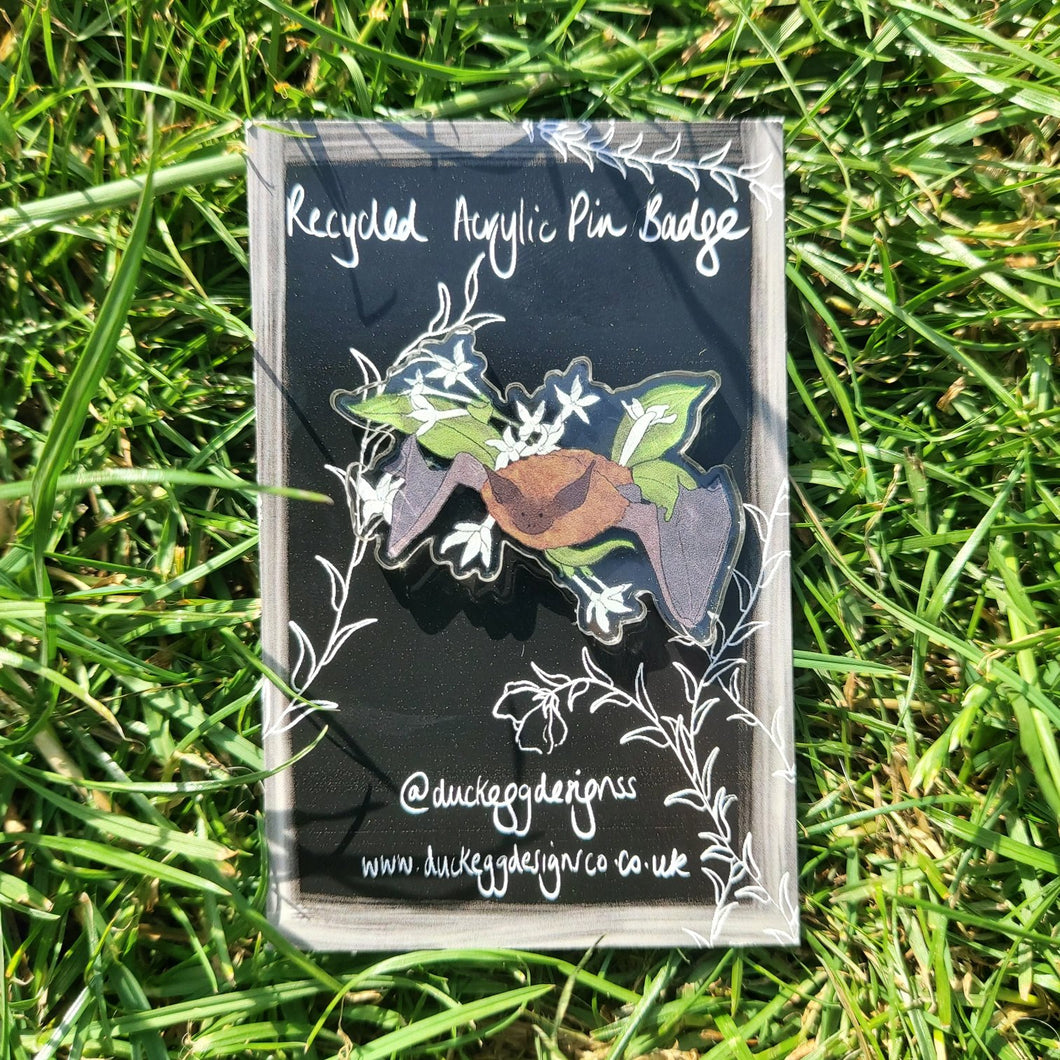 In the middle of a grassy background you can see a clear acrylic pin badge featuring a brown and grey pipistrelle bat surrounded by jasmine. The badge is on a black backing card which has a white design featuring the words ‘Recycled Acrylic Pin Badge’ and a white brushstroke border as well as leafy vines.