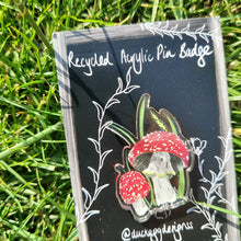 Load image into Gallery viewer, A close up of the red and white fly agaric badge on its black and white backing card. Behind the backing card you can see a green grassy background.
