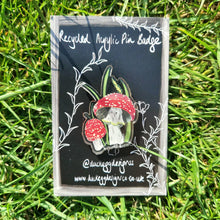 Load image into Gallery viewer, A black backing card featuring a white design sits in front of a green grassy background. In the centre of the backing card is a shiny acrylic pin badge featuring a fly agaric and wild grasses design. 
