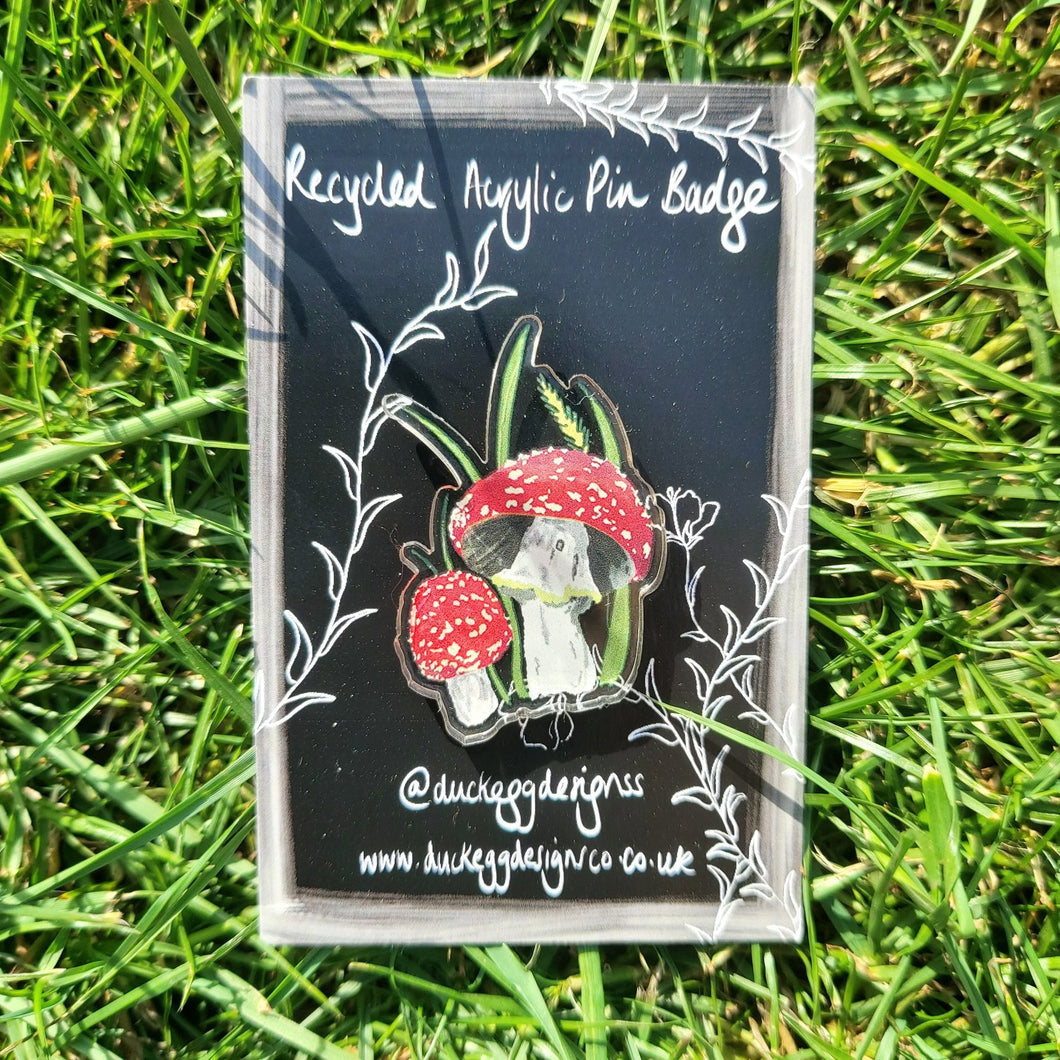 A black backing card featuring a white design sits in front of a green grassy background. In the centre of the backing card is a shiny acrylic pin badge featuring a fly agaric and wild grasses design. 