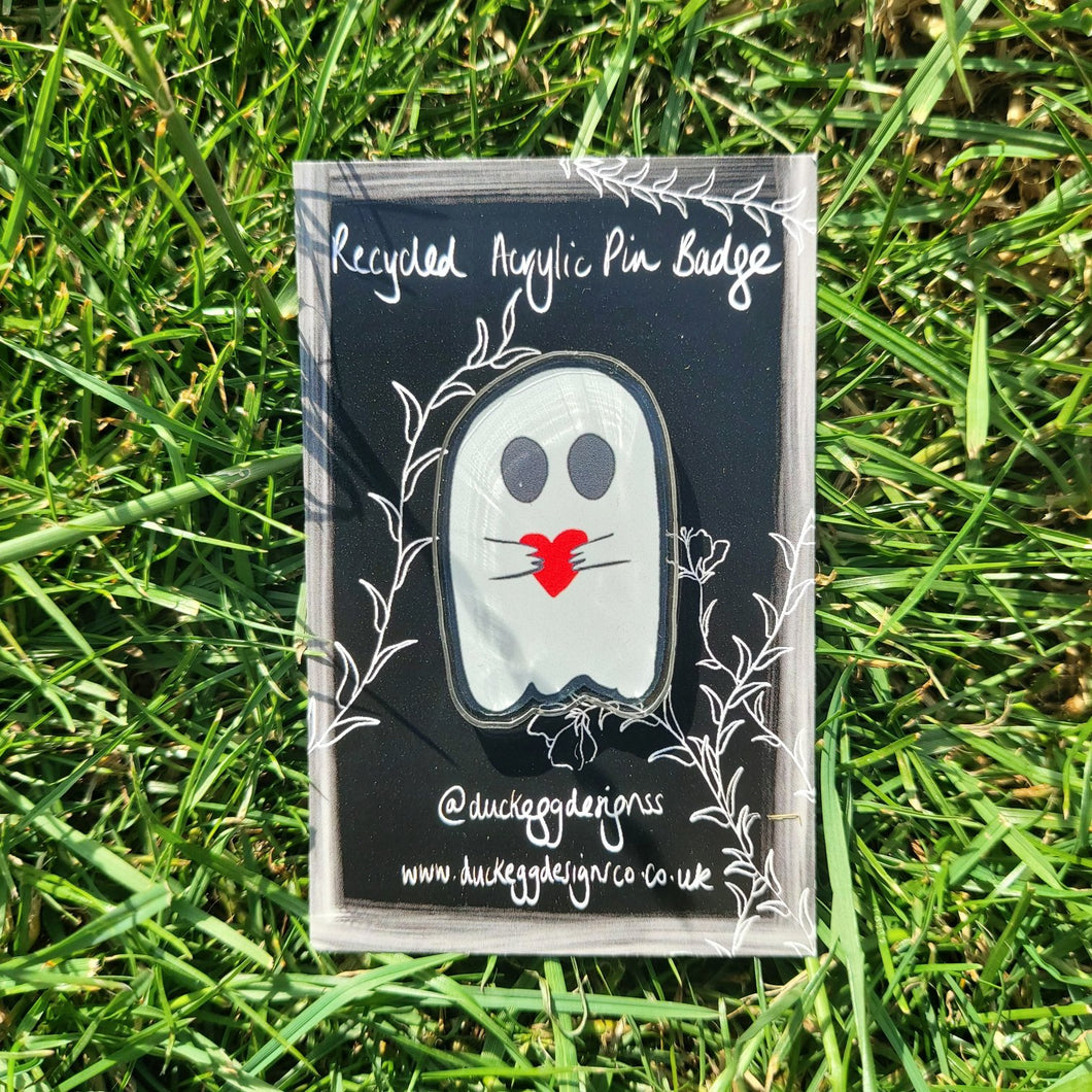 In the middle of a green grassy background you can see a black and white backing card with the words ‘ Recycled Acrylic Pin Badge’ in white handwriting. In the centre of the backing card you can see a shiny ghost pin badge featuring a white gloss with black eyes holding a red heart. 