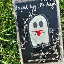 Load image into Gallery viewer, A close up of the ghost acrylic pin badge featuring a black outline around a white ghost with black eyes holding a red heart. The ghost sits on a black and white backing card behind which you can see green grass.
