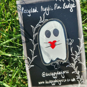 A close up of the ghost acrylic pin badge featuring a black outline around a white ghost with black eyes holding a red heart. The ghost sits on a black and white backing card behind which you can see green grass.