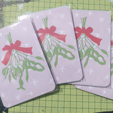 Load image into Gallery viewer, Four packs of Christmas postcards across a green cutting mat. You can see the top postcard in the pack which is a pale pink design featuring white snowflakes and a bunch of mistletoe tied by a red ribbon hanging down the middle.
