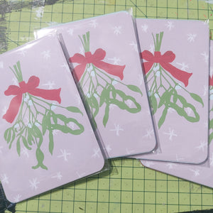 Four packs of Christmas postcards across a green cutting mat. You can see the top postcard in the pack which is a pale pink design featuring white snowflakes and a bunch of mistletoe tied by a red ribbon hanging down the middle.