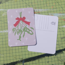 Load image into Gallery viewer, A pink postcard sits on a green cutting mat, featuring a bunch of green mistletoe tied by a red ribbon hanging down the middle. Behind the mistletoe you can see white snowflake shapes. Under the top postcard you can see an upside down postcard featuring a white back with lines to put the address in, a box for the stamp and a duck egg designs logo.
