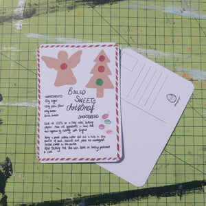 Front and back of a postcard shown on a green cutting mat. The front shows a handwritten boiled sweets Christmas shortbread recipe bordered by red and white candy cane, with angel and Christmas tree biscuits on the card. 