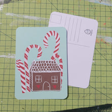 Load image into Gallery viewer, Front and back of a postcard are visible on a green cutting mat. Front of the postcard shows a gingerbread house with red and white icing under some tall red and white candy canes on a peppermint green background. The back of the postcard is white, featuring lines for the address and a square for the stamp.
