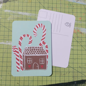 Front and back of a postcard are visible on a green cutting mat. Front of the postcard shows a gingerbread house with red and white icing under some tall red and white candy canes on a peppermint green background. The back of the postcard is white, featuring lines for the address and a square for the stamp.