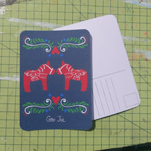 Load image into Gallery viewer, Front and back of a postcard are visible on a green cutting mat. The front is dark blue and features red dala horses with the words ‘God Jul’ underneath in white. The back of the postcard is white and has lines for the address, a box for the stamp and a duck egg designs logo.
