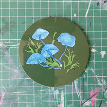 Load image into Gallery viewer, A dark green coaster featuring electric blue pixie parasol coasters with some green moss. Behind the coaster you can see a green cutting mat with paint streaks.
