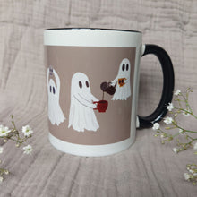 Load image into Gallery viewer, A white and warm grey mug featuring ghost with a black interior and handle sits in front of a greyish lilac material in between two sprigs of babys breath. The two ghosts you can see the most are sharing a pot of coffee - with one of the ghost floating above the other.
