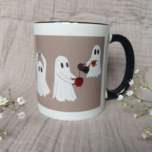 A white and warm grey mug featuring ghost with a black interior and handle sits in front of a greyish lilac material in between two sprigs of babys breath. The two ghosts you can see the most are sharing a pot of coffee - with one of the ghost floating above the other.