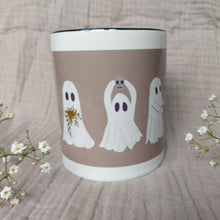 Load image into Gallery viewer, A white and warm grey mug featuring ghost with a black interior sits in front of a greyish lilac material in between two sprigs of babys breath. The two ghosts you can see clearly hold a plant pot with a green trailing plant, and a mini skull.
