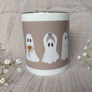 A white and warm grey mug featuring ghost with a black interior sits in front of a greyish lilac material in between two sprigs of babys breath. The two ghosts you can see clearly hold a plant pot with a green trailing plant, and a mini skull.