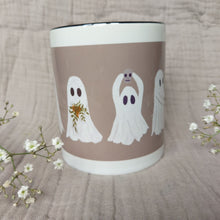 Load image into Gallery viewer, A white and warm grey mug featuring ghost with a black interior sits in front of a greyish lilac material in between two sprigs of babys breath. The two ghosts you can see clearly hold a plant pot with a green trailing plant, and a mini skull.
