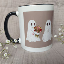 Load image into Gallery viewer, A mug featuring ghost with a black handle and interior sits in front of a greyish lilac material in between two sprigs of babys breath. The ghost you can see the most of carries a bunch of flowers.
