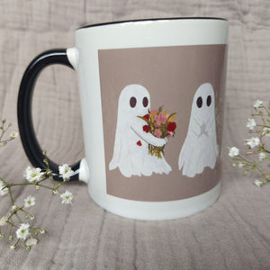 A mug featuring ghost with a black handle and interior sits in front of a greyish lilac material in between two sprigs of babys breath. The ghost you can see the most of carries a bunch of flowers.