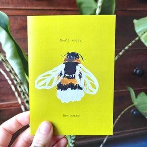 Warm yellow card featuring a hand drawn bee and the words 'Don't worry Bee happy'. Behind the card is a stripy stemmed leafy houseplant, behind which is a mahogany chest of draws.
