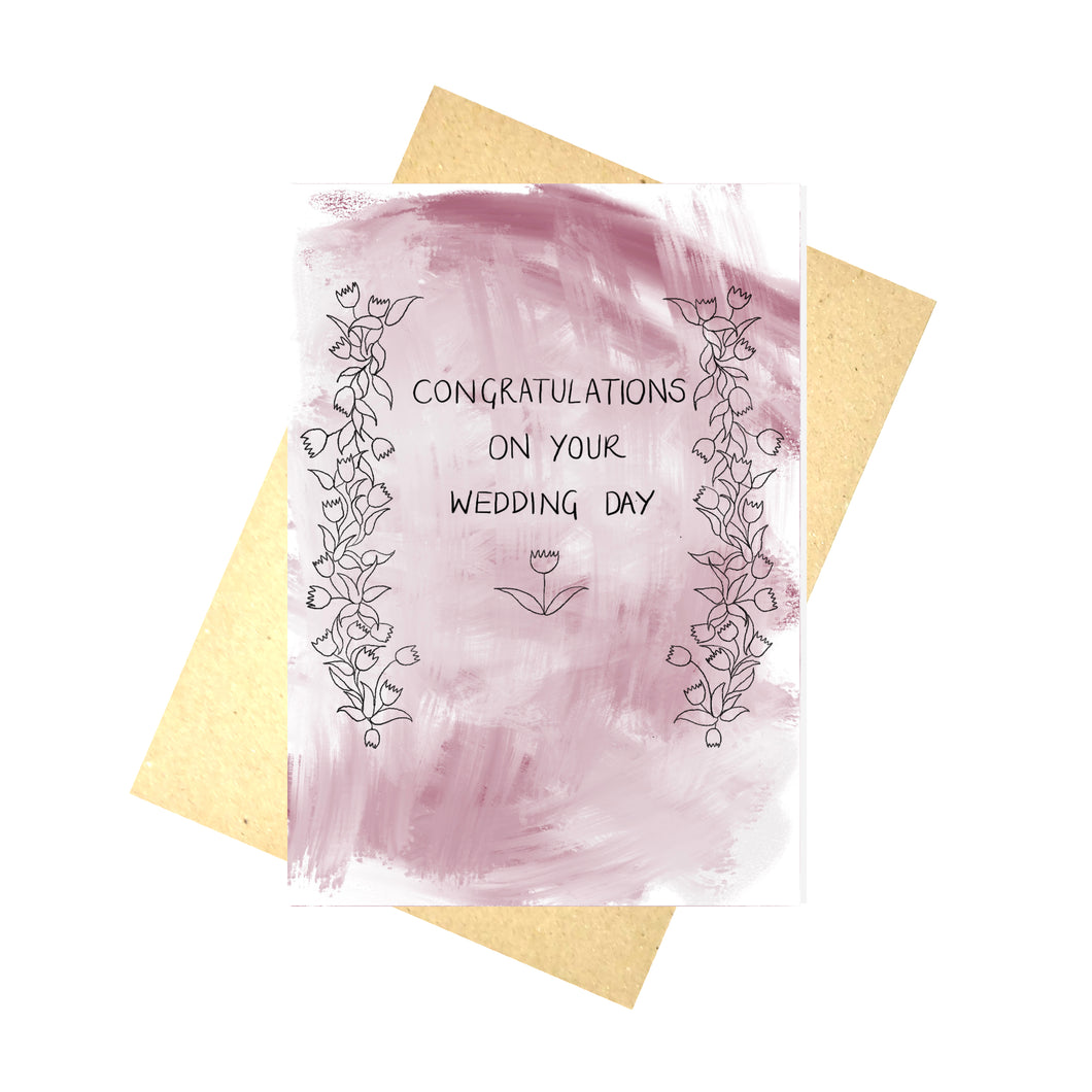 Simple wedding card with a textured pastel red/pink background. Black floral vines line the left and right of the card, with a flower with leaves visible below the words 'Congratulations On Your Wedding Day'. Behind the card is a brown envelope, behind which is a white background.