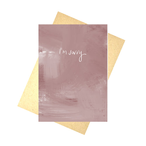 A dusky pink card sits on a brown recycled envelope in front of a white background. The card has the words 'I'm sorry' in white handwriting