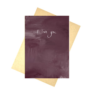 A deep plummy purple tonal card sits on a recycled brown envelope in front of a white background. The card has the words 'I love you' in white handwriting in the middle.