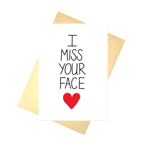 A white card with the words 'I MISS YOUR FACE' down the middle, below which is a red heart. Behind the card is a brown envelope behind which is a white background. 