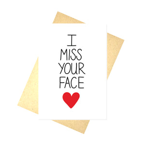 A white card with the words 'I MISS YOUR FACE' down the middle, below which is a red heart. Behind the card is a brown envelope behind which is a white background. 