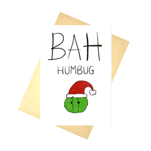 White christmas card featuring the words BAH HUMBUG in black block handwriting above a little green brussel sprout wearing a red and black santa hat. Behind the card is a brown recycled envelope in front of a white background.