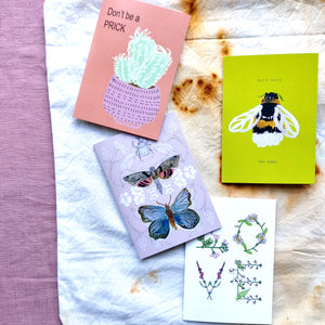 Four cards in front of rust dyed fabric with a strip of lilac to the left. Top left is the orange 'Don't be a PRICK 'card, to the right of it is the yellow Bee Happy card featuring a bumblebee. Underneath the orange card is pale off pink card featuring a rhino beetle, moth and butterfly, under which is a white card featuring LOVE written in flowers.