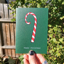 Load image into Gallery viewer, A green card featuring a white and red candy cane with white writing below that says &#39;Happy Christmas&#39;. The card is being held in front of a sunny garden with a wooded fence by a hand with black nail polish.
