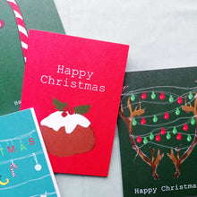 Load image into Gallery viewer, A group of four cards arranged in a pile - a red card with a christmas pudding is most visible and has the words &#39;Happy Christmas&#39; in white. Under the red card to the left is a dark green card featuring reindeer antlers with christmas lights strung between them. To the left of those cards is a blue christmas card and below the red card is a dark green card with a white and red candy cane. Behind the cards is a pale green fabric background.
