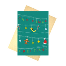 Load image into Gallery viewer, A blue card with white lines across it, from which the letters for the works &#39;Christmas Greetings&#39; hand in alternating green and red letters. Hanging across the middle two rows are a mix of red and white candy canes, green, blue and yellow baubles, a yellow elf shoe and a santa hat. Behind the card is a brown envelope, behind which is a white background.
