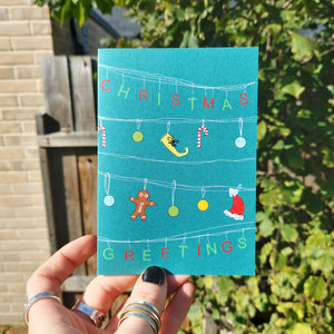 A blue card with white lines across it, from which the letters for the works 'Christmas Greetings' hand in alternating green and red letters. Hanging across the middle two rows are a mix of red and white candy canes, green, blue and yellow baubles, a yellow elf shoe and a santa hat.  The card is held by a hand with black nail polish and features a sunny garden with a wooden fence behind it.
