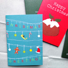 Load image into Gallery viewer,  A blue card with white lines across it, from which the letters for the works &#39;Christmas Greetings&#39; hand in alternating green and red letters. Hanging across the middle two rows are a mix of red and white candy canes, green, blue and yellow baubles, a yellow elf shoe and a santa hat.  Behind the card is a red card featuring a christmas pudding, behind which are two green cards, and a pale green background fabric.
