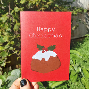 A red card featuring a simple christmas pudding illustration. Above the pudding are the words 'Happy Christmas' in contrasting white. The card is held by a hand with black nail polish, and behind it is a sunny green garden.