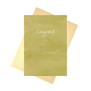 A tonal deep mustard yellow card sits in front of a recycled brown paper envelope in front of a white background. The card says 'Congrats!' in white handwriting across the middle. 
