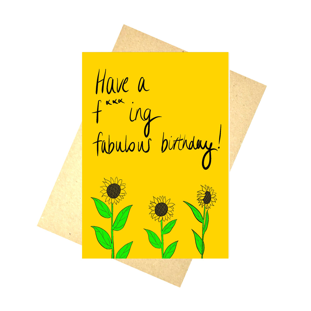 Warm yellow card with the words 'Have a f***ing fabulous birthday!' in black handwriting above some sunflowers growing up from the bottom of the card. Behind the card you can see a brown envelope in front of a white background.
