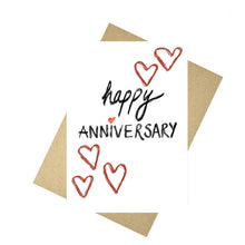 Load image into Gallery viewer, White card featuring the words happy anniversary in black textured handwriting with a red love heart for the i in anniversary. Above and below the writing are a few red hearts in textured brush strokes. Behind the card a brown envelope is visible, behind which is a white background.
