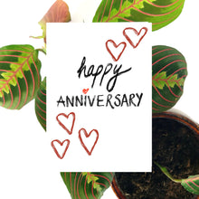 Load image into Gallery viewer, White card featuring the words happy anniversary in black textured handwriting with a red love heart for the i in anniversary. Above and below the writing are a few red hearts in textured brush strokes. Behind the card a brown envelope is visible, behind which is a prayer plant on a white background.
